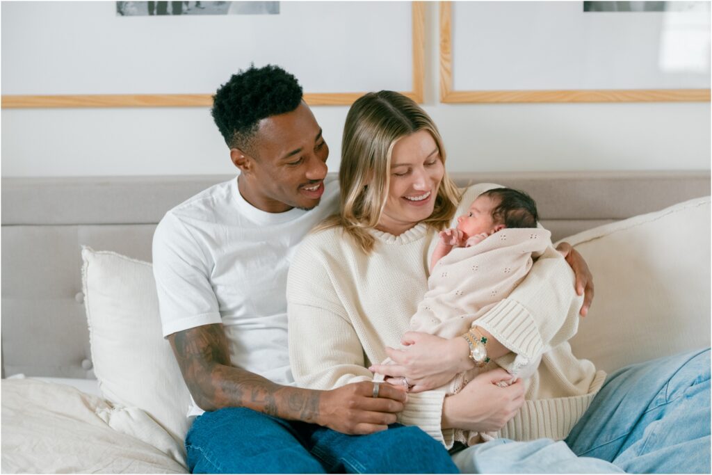 Family with newborn baby sitting on a bed in a bright room.