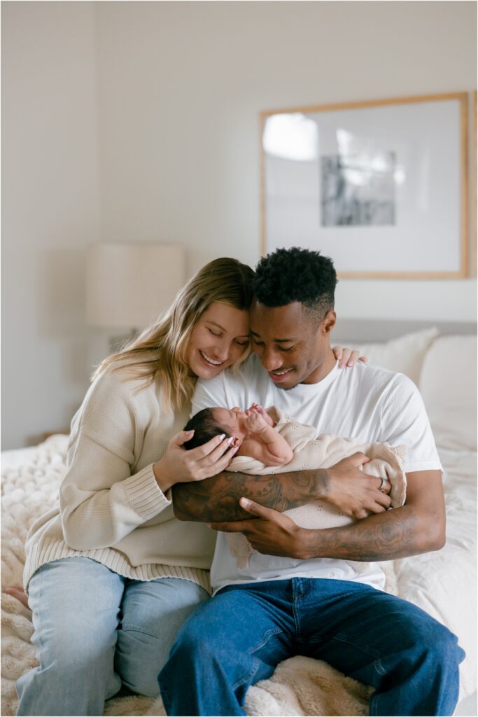 Parents hold their newborn baby on a bed in a cozy, well-lit room.
