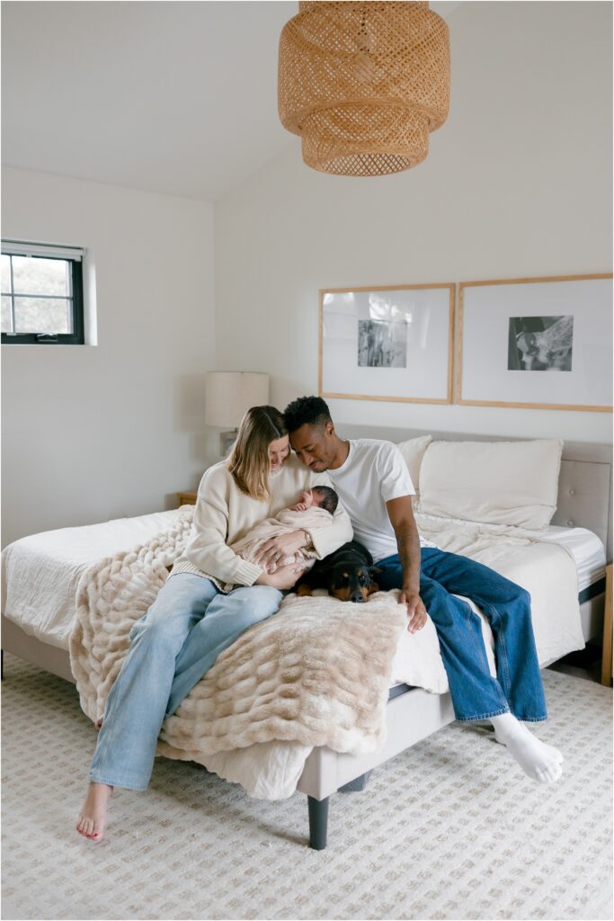 Family with newborn and doberman dog baby sitting on bed in a bright bedroom with minimal decor.